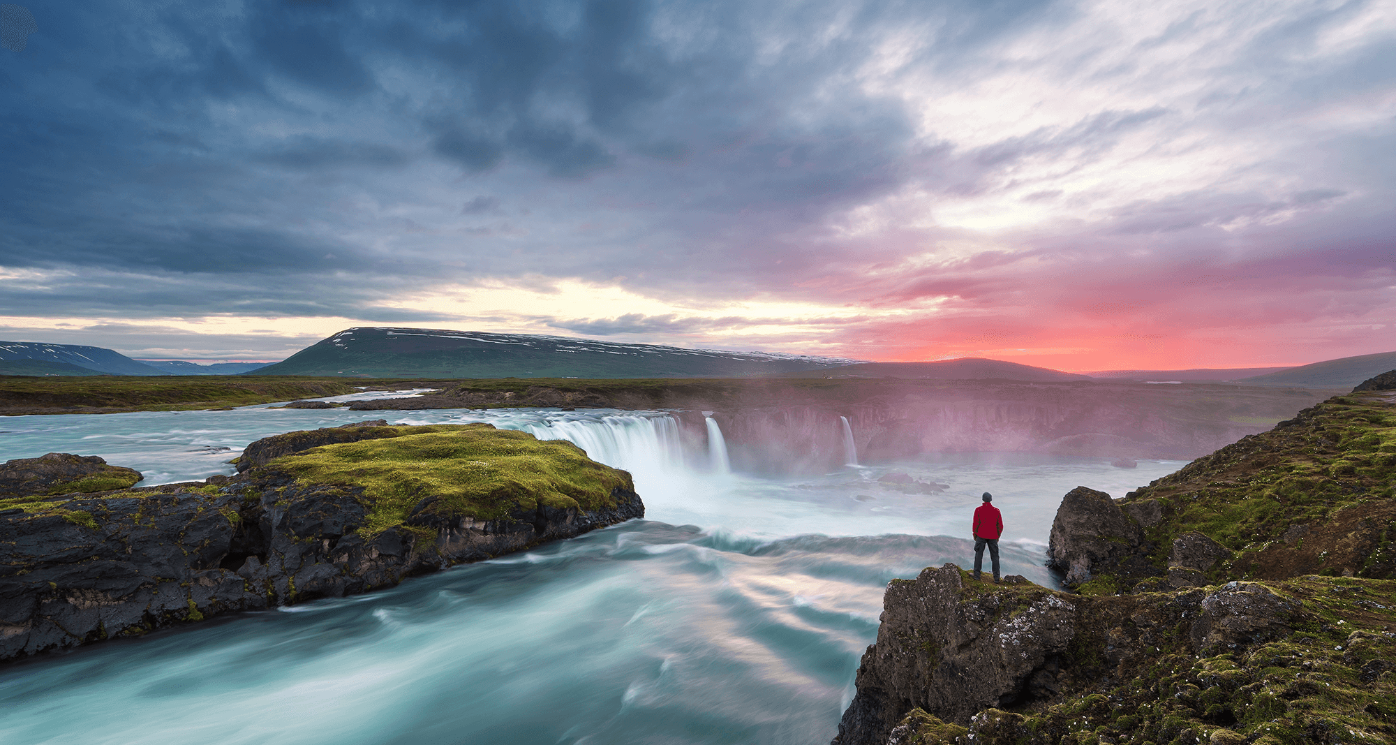 Ein Mann steht vor einer Landschaft mit Wasserfall in der Abenddämmerung. Das Bild symbolisiert unseren Claim Think. Create. Change.